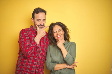 İzole sarı arka plan üzerinde güzel orta yaş çift çapraz silah ve el çene üzerinde yükseltilmiş ile gülümseyen kamera emin bakıyor. Pozitif düşünme.