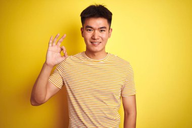Genç asyalı Çinli adam izole sarı arka plan üzerinde duran t-shirt giyen olumlu el ve parmakları ile ok işareti yapıyor gülümseyerek. Başarılı ifade.