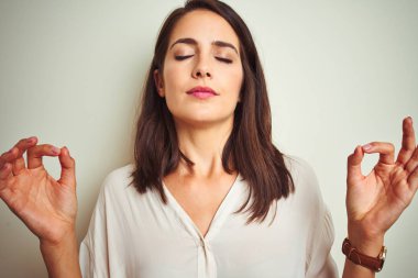 Beyaz izole arka plan üzerinde duran rahat gömlek giyen genç güzel kadın rahat ve gözleri parmakları ile meditasyon jest yapıyor kapalı gülümseyerek. Yoga konsepti.