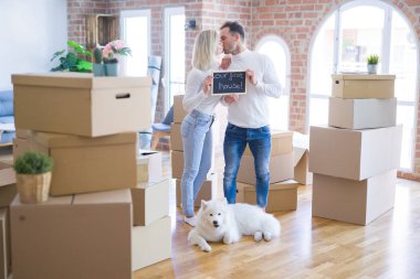 Karton kutular etrafında yeni evde öpüşme mesajı ile tahta tutan ayakta köpek ayakta genç güzel çift