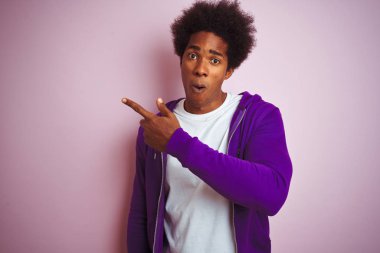 İzole pembe arka plan üzerinde duran mor sweatshirt giyen genç afro-amerikan adam yan parmak ile işaret şaşırttı, açık ağız hayrete ifade.