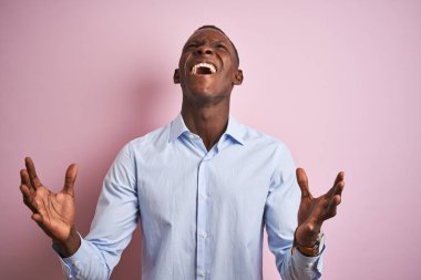 Mavi zarif gömlek giyen Afro-amerikan adam izole pembe arka plan üzerinde duran deli ve deli bağırarak ve agresif ifade ve kollar kaldırdı ile bağırıyor. Hayal kırıklığı kavramı.