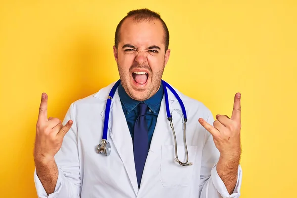 Young doctor man wearing coat and stethoscope standing over isolated yellow background shouting with crazy expression doing rock symbol with hands up. Music star. Heavy concept.