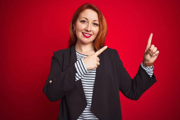 Beautiful redhead business woman wearing elegant jacket over isolated red background smiling and looking at the camera pointing with two hands and fingers to the side.