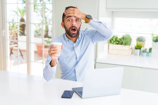 Handsome hispanic man working using computer laptop and drinking a cup of coffee stressed with hand on head, shocked with shame and surprise face, angry and frustrated. Fear and upset for mistake.