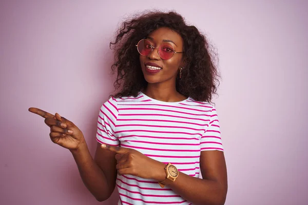 Young african american woman wearing t-shirt and sunglasses over isolated pink background smiling and looking at the camera pointing with two hands and fingers to the side.