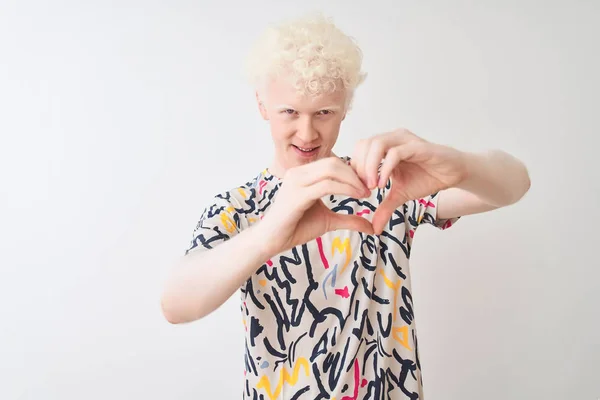 Young albino blond man wearing colorful t-shirt standing over isolated white background smiling in love showing heart symbol and shape with hands. Romantic concept.