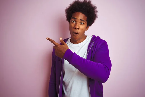 年轻的非洲裔美国男子穿着紫色运动衫站在孤立的粉红色背景惊讶用手指指着一边 张开嘴惊讶的表情 — 图库照片