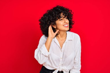 İzole kırmızı arka plan üzerinde beyaz rahat gömlek giyen kıvırcık saçlı genç arap kadın kulak üzerinde el ile gülümseyerek söylenti veya dedikodu bir işitme dinlerken. Sağırlık kavramı.