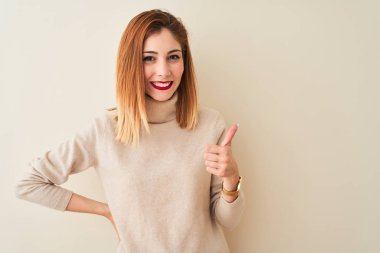 Kırmızı saçlı kadın zarif boğazlı kazak giyen izole beyaz arka plan üzerinde duran el ile jest kadar mutlu başparmak yapıyor. Başarı gösteren kameraya bakarak ifadeyi onaylama.