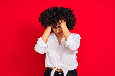 Kıvırcık saçlı genç arap kadın izole kırmızı arka plan üzerinde beyaz rahat gömlek giyen baş ağrısı umutsuz muzdarip ve stresli çünkü ağrı ve migren. Eller baş üstünde.