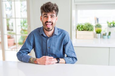 Beyaz masanın üzerinde oturan rahat gömlek giyen genç adam komik ifade ile mutlu dil dışarı yapışmasını. Duygu kavramı.