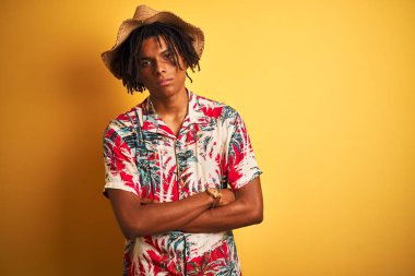 Izole sarı arka plan şüpheci ve sinir üzerinde çiçek gömlek ve şapka giyen dreadlocks ile Afro Amerikan adam, çapraz kolları ile yüzünde onaylamayan ifade. Negatif kişi.