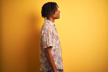 Yan bakarak izole sarı arka plan üzerinde çiçek gömlek giyen tatilde dreadlocks ile Afro adam, kendine güvenen gülümseme ile doğal yüzü ile profil poz rahatla.