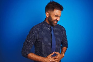 Genç Hintli işadamı zarif gömlek ve kravat mide üzerinde el ile izole mavi arka plan üzerinde duran giyen hazımsızlık, ağrılı hastalık kötü hissediyorum. Ağrı kavramı.