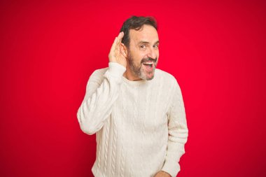 Izole kırmızı arka plan üzerinde gri saçlı Yakışıklı orta yaş yaşlı adam söylenti veya dedikodu bir işitme dinlerken kulak üzerinde el ile gülümseyerek. Sağırlık kavramı.