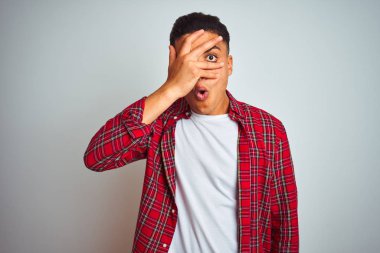 Izole beyaz arka plan üzerinde duran kırmızı gömlek giyen genç Brezilyalı adam şok yüz ve gözleri el ile kaplayan gözetleme, utanmış ifade ile parmaklarından bakarak.