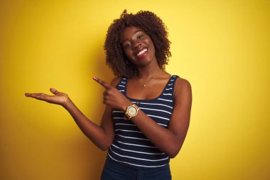 Genç afrikalı afro kadın izole sarı arka plan üzerinde çizgili t-shirt giyen hayran ve el ile sunarken kameraya gülümseyerek ve parmak ile işaret.