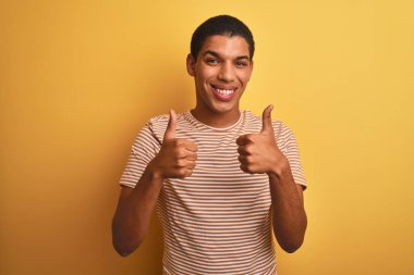 İzole sarı arka plan başarı işareti el ile olumlu jest yapıyor çizgili t-shirt giyen genç yakışıklı Arap adam, gülümseyerek ve mutlu başparmak kadar. Neşeli ifade ve kazanan jest.