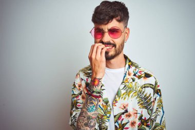 Tatilde yazlık gömlek güneş gözlüğü giyen dövmeli adam izole beyaz arka plan üzerinde stresli ve ağız tırnak ısırma elleri ile sinir görünümlü. Anksiyete sorunu.