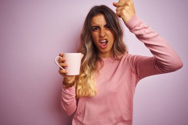 Genç güzel kadın pembe izole arka plan üzerinde kahve fincan tutan kızgın ve sinirli öfke, deli ve yükseltilmiş el, öfke kavramı ile bağırarak