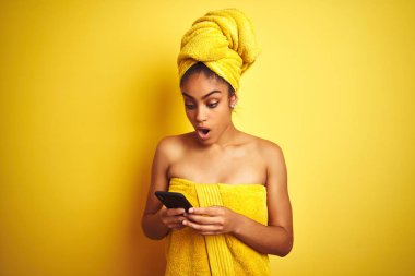 Afro kadın izole sarı arka plan üzerinde smatrphone kullanarak duş tan sonra havlu giyen sürpriz bir yüz ile şok korkmuş, korku ve korku ifade ile heyecanlı