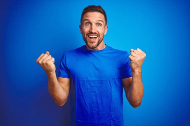 Genç yakışıklı adam mavi izole arka plan üzerinde rahat t-shirt giyen sürpriz ve kollarını kaldırdı ve açık gözleri ile başarı için şaşırttı kutluyor. Kazanan kavramı.