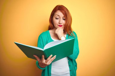 Genç kızıl saçlı öğrenci kadın sarı izole arka plan üzerinde yeşil kitap okuma ciddi yüz soru hakkında düşünme, çok karışık bir fikir