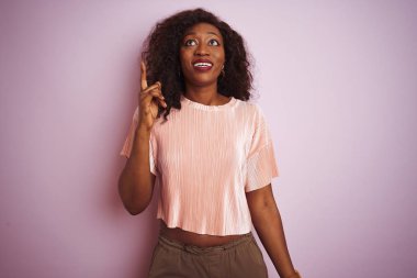 Genç afro-amerikan kadın izole pembe arka plan üzerinde duran t-shirt giyen başarılı bir fikir ile parmak işaret. Çıktı ve mutlu. Bir numara.