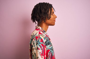 Yan bakarak izole pembe arka plan üzerinde çiçek gömlek giyen tatilde dreadlocks ile Afro adam, kendine güvenen gülümseme ile doğal yüzü ile profil poz rahatla.