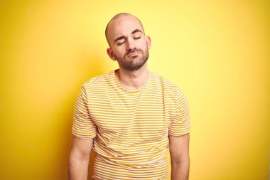 Sakallı genç kel adam sarı izole arka plan üzerinde rahat çizgili t-shirt giyen uykulu ve yorgun görünümlü, yorgunluk ve akşamdan kalma, sabah tembel gözleri için yorgun.