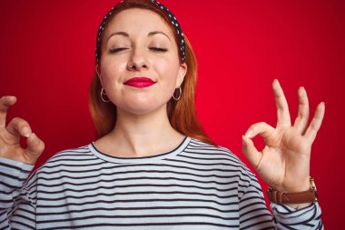 İzole kırmızı arka plan üzerinde duran çizgili lacivert t-shirt giyen güzel kızıl saçlı kadın rahatlayın ve parmakları ile meditasyon jest yapıyor kapalı gözleri ile gülümseyerek. Yoga konsepti.