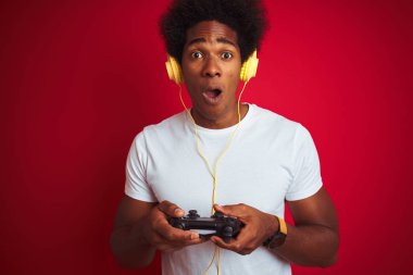 Afro oyun adam izole kırmızı arka plan üzerinde joystick kulaklık kullanarak video oyunu oynarken sürpriz bir yüz ile şok korkmuş, korku ve korku ifadesi ile heyecanlı
