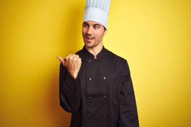 Üniforma ve şapka giyen genç şef adam izole sarı arka plan üzerinde duran mutlu yüz görünümlü ve başparmak ile yan işaret gülümseyerek.