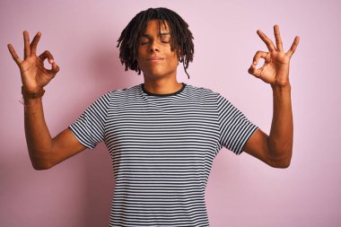 Izole pembe arka plan üzerinde duran lacivert çizgili t-shirt giyen dreadlocks ile Afro adam rahatlayın ve parmakları ile meditasyon jest yapıyor kapalı gözleri ile gülümseyerek. Yoga konsepti.