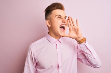 Genç yakışıklı işadamı izole pembe arka plan üzerinde duran zarif gömlek giyen bağırarak ve ağızda el ile yan yüksek sesle bağırıyor. İletişim kavramı.