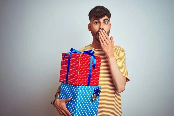 Young man with tattoo holding birthday gifts standing over isolated white background cover mouth with hand shocked with shame for mistake, expression of fear, scared in silence, secret concept