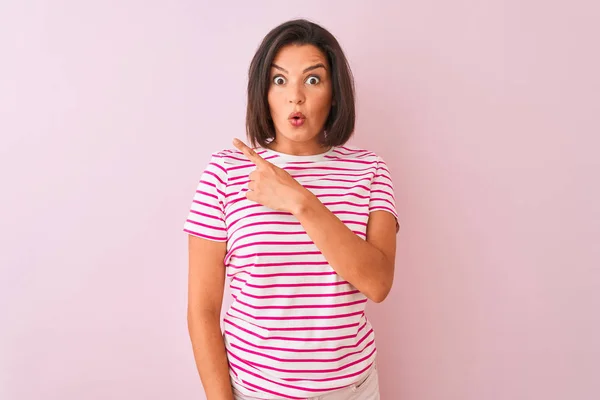 年轻美丽的女人穿着条纹T恤站在孤立的粉红色背景惊讶用手指指着一边 张开嘴惊讶的表情 — 图库照片