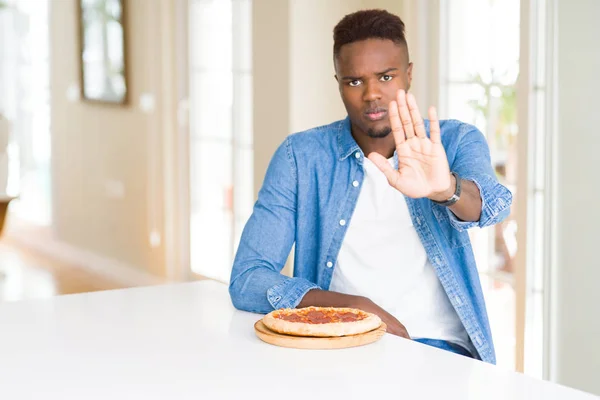 African american man eating pepperoni pizza at home with open hand doing stop sign with serious and confident expression, defense gesture