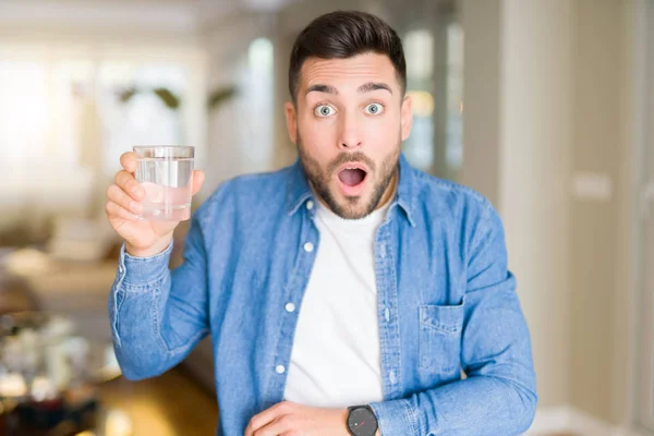 自宅で水のコップを飲む若いハンサムな男は 驚きの顔でショックを受け 恐怖の表情で恐怖と興奮 — ストック写真