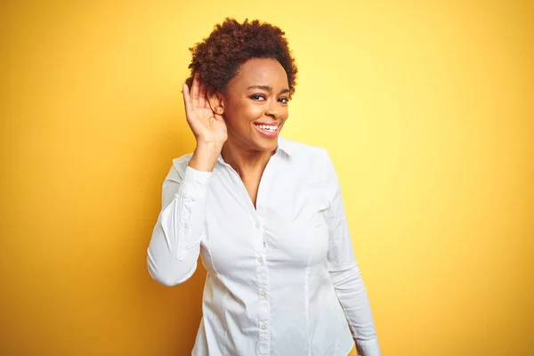 African american business woman over isolated yellow background smiling with hand over ear listening an hearing to rumor or gossip. Deafness concept.