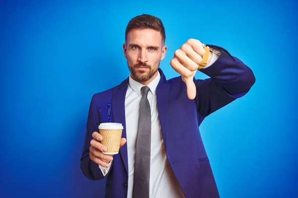 Young handsome business man drinking a coffee on a paper cup over blue isolated background with angry face, negative sign showing dislike with thumbs down, rejection concept