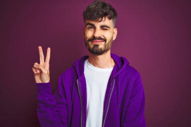 Çizgili polo giyen dövmeli genç adam izole sarı arka plan üzerinde duran mutlu yüz zafer işareti yapıyor kameraya göz kırparak gülümseyerek. İki numara.