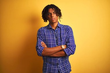 Izole sarı arka plan şüpheci ve sinir üzerinde duran rahat gömlek giyen dreadlocks ile Afro adam, çapraz kolları ile yüzünde ifade onaylamayan. Negatif kişi.