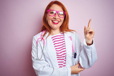 Genç kızıl saçlı doktor kadın yüzünde büyük bir gülümseme ile izole pembe arka plan üzerinde duran stetoskop kullanarak, kameraya bakarak yan el ve parmak ile işaret.