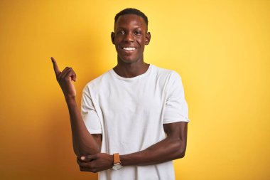 Beyaz tişört giyen genç afro-amerikan adam yüzünde büyük bir gülümseme ile izole sarı arka plan üzerinde duran, kameraya bakarak yan el ve parmak ile işaret.