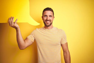 Young man holding empty speech bubble for message over yellow isolated background with a happy face standing and smiling with a confident smile showing teeth
