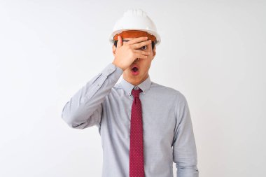 Chinese architect man wearing tie and helmet standing over isolated white background peeking in shock covering face and eyes with hand, looking through fingers with embarrassed expression.