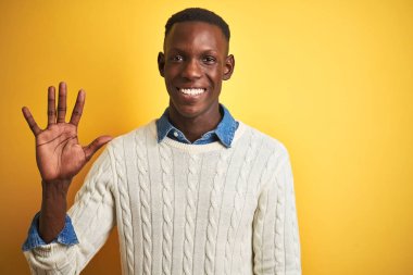 Afro-amerikan adam denim gömlek ve izole sarı arka plan üzerinde beyaz kazak giyen gösteren ve parmakları ile işaret ederken emin ve mutlu gülümseyerek beş numara.