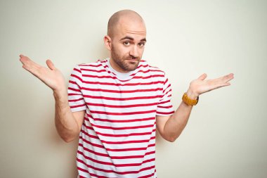 Sakallı genç kel adam beyaz izole arka plan üzerinde rahat çizgili kırmızı t-shirt giyen habersiz ve kollar ve eller kaldırdı ile karışık ifade. Şüphe kavramı.